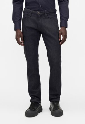 Homme portant un jean slim foncé, des baskets noires et une chemise sombre à manches longues, les mains reposant le long du corps.