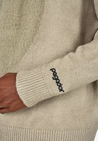 Beige pulóver, textúrált zöld díszítéssel, bordás mandzsettákkal és fekete „pegador” logóval hímzéssel az ujjon. Puha, kötött anyag.