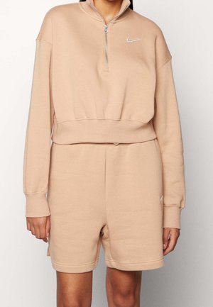 Femme portant un sweat Nike beige à fermeture éclair quart et un short assorti, debout devant un fond blanc uni.