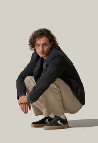 Blazer nero, camicia grigia, pantaloni beige e sneakers nere con suola bianca e color gomma. Fashionato in posizione accovacciata, su uno sfondo neutro.