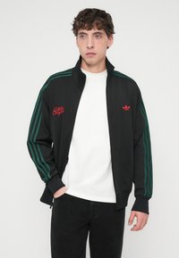 Sort løbetrøje med grønne striber og rødt broderet logo. Har lynlåslukning foran og en opretstående krave, lavet af et glat materiale.