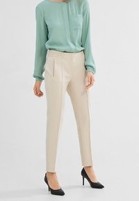 Helle grüne Bluse mit einer Tasche; cremefarbene Slim-Fit-Hose; schwarze Hochzeitschuhe; glattes Material und maßgeschneiderter Schnitt.