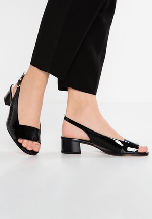 Fødder iført sorte slingback-sandaler i laklæder med lave blokhæle og åbne tæer, kombineret med sorte korte bukser.