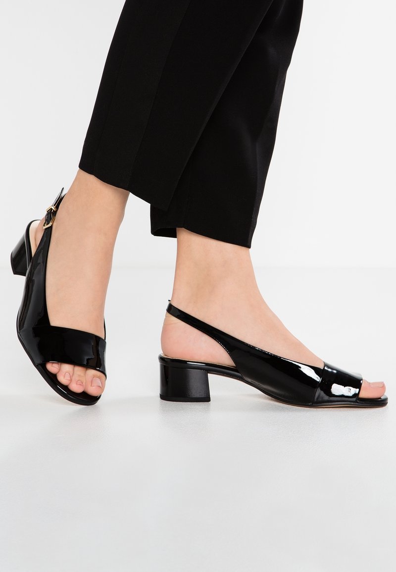 Pieds portant des sandales slingback en cuir verni noir avec des talons blocs bas et des orteils ouverts, associées à un pantalon noir raccourci.