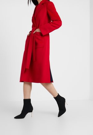 Mujer caminando con un abrigo largo rojo brillante con cinturón y grandes bolsillos parche, llevando botines negros de tacón alto y punta.