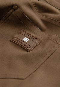 Tissu marron en gros plan avec une poche cousue et un patch rectangulaire marron présentant un petit emblème carré blanc au centre.