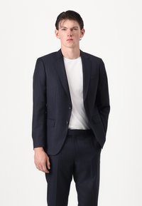 Nuori mies, joka pukeutunut tummansiniseen liituraitapukuun ja valkoiseen t-paitaan, seisoo käsi taskussa yksinkertaisen valkoisen taustan edessä.