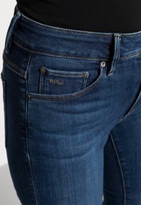 Jeans de mezclilla azul oscuro con una textura suave, que cuentan con un solo bolsillo frontal, herrajes de latón y detalles de costura, etiquetados como "RAW".