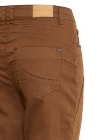 Jean en denim marron en gros plan montrant la poche arrière avec rivets et patch de marque en cuir Pulz Jeans sur la ceinture.