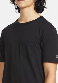 Sort bomulds t-shirt med rund hals, korte ærmer og en lomme på venstre bryst, samt en logo detalje på højre ærme.