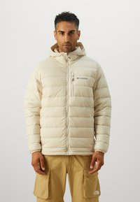 Beige puffer jacket with a hood, quilted design, front zipper, and logo on the left chest. Worn with tan cargo pants.