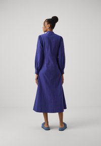 Abito lungo a maniche lunghe, fino alla caviglia, in lino viola scuro con decorazioni ricamate sulle maniche e dettagli con bottoni. Indossato con scarpe blu da infilare.