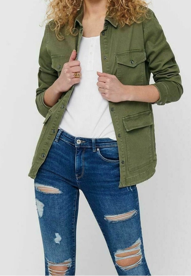 Giacca verde oliva con bottoni, con due tasche sul petto, indossata sopra una maglietta bianca e jeans blu strappati, con multiple abrasioni.