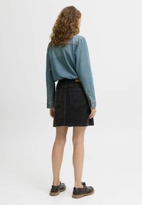 Kvinna med lockigt hår som bär en blå denimjacka, svart denimkjol och svarta skor, stående med ryggen mot kameran.