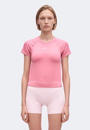 Femme debout, face à l'avant, portant un t-shirt rose à manches courtes Nike et un short rose clair Nike sur un fond uni.