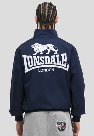 Persoon met gevlochten haar draagt een marineblauw jack met wit Lonsdale London-logo en grijze broek met Britse vlagpatch op de zak.