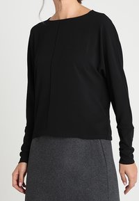 Svart långärmad topp med rundad halsringning, texturerad tyg, avslappnad passform och synlig sömdetalj, tillsammans med en grå pennkjol.