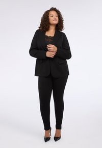 Černý propracovaný blejzr s odpovídajícími přiléhavými kalhotami, které mají elegantní design, hladkou texturu a špičaté černé podpatky. Viditelný top s krajkovými doplňky.