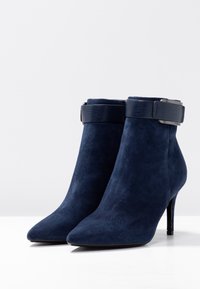 Navy blauwe suède enkellaarsjes met een puntige neus, hoge hak en een gestructureerde band rond de enkel, voorzien van een metalen accent.