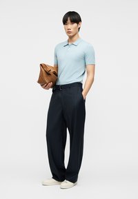 Νεαρός άνδρας με ανοιχτό μπλε πόλο πουκάμισο, μαύρο παντελόνι με φαρδιές ρίγες, λευκά αθλητικά παπούτσια, κρατώντας καφέ δερμάτινη τσάντα, στέκεται μπροστά σε απλό φόντο.