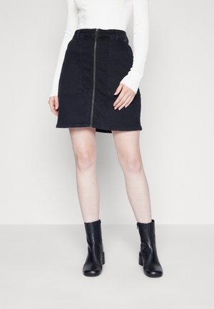 Femme portant une jupe noire zippée sur le devant, un haut blanc à manches longues et des bottines noires, debout devant un fond clair uni.