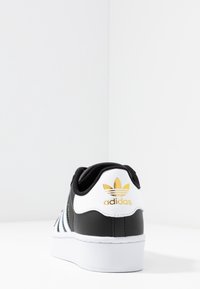 Mustat ja valkoiset Adidas-tennarit, joissa on kultainen logo, nahkamateriaali, matala profiili, teksturoitu kankainen kieli ja mintunvihreät raidat.