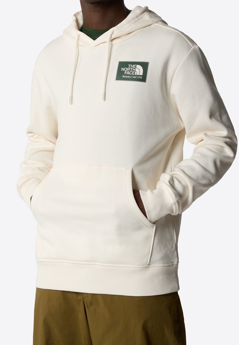 Cremefarbener Kapuzenpullover mit einer Fronttasche, gerippten Bündchen und Saum. Besitzt ein grünes Markenpatch auf der oberen linken Brustseite. Weicher Stoff.
