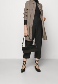 Kvinna iklädd en beige lång jacka, svarta cropped byxor, svarta klackar med remmar, hållande en svart handväska med guldkedjedetaljer.