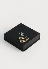 Cercei lungi de culoare aurie, cu pietre clare, de formă rectangulară, prezentate într-o cutie neagră elegantă, cu un logo argintiu pe capac.