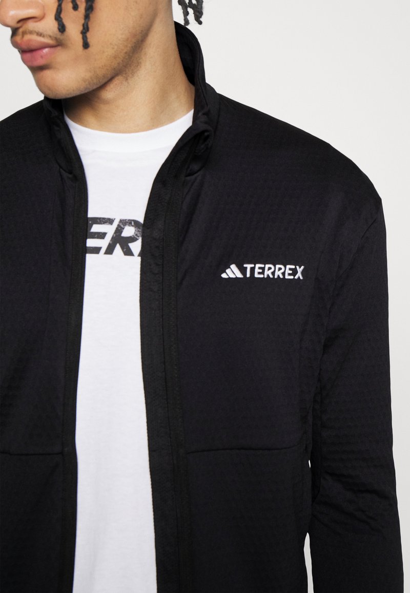 Man in een zwart jack met rits en het witte "TERREX" logo, over een wit shirt met gedeeltelijk zichtbare zwarte tekst.
