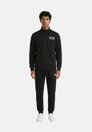Giovane uomo in piedi che indossa una giacca nera con zip e logo sul petto, pantaloni neri abbinati e scarpe da ginnastica bianche su uno sfondo semplice.
