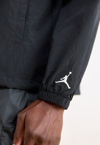 Μαύρο νάιλον μπουφάν με ελαστικούς καρπούς. Διαθέτει το λευκό λογότυπο Jumpman στο αριστερό μανίκι. Λείος υφασμάτινος σχεδιασμός, ελαφρύς.