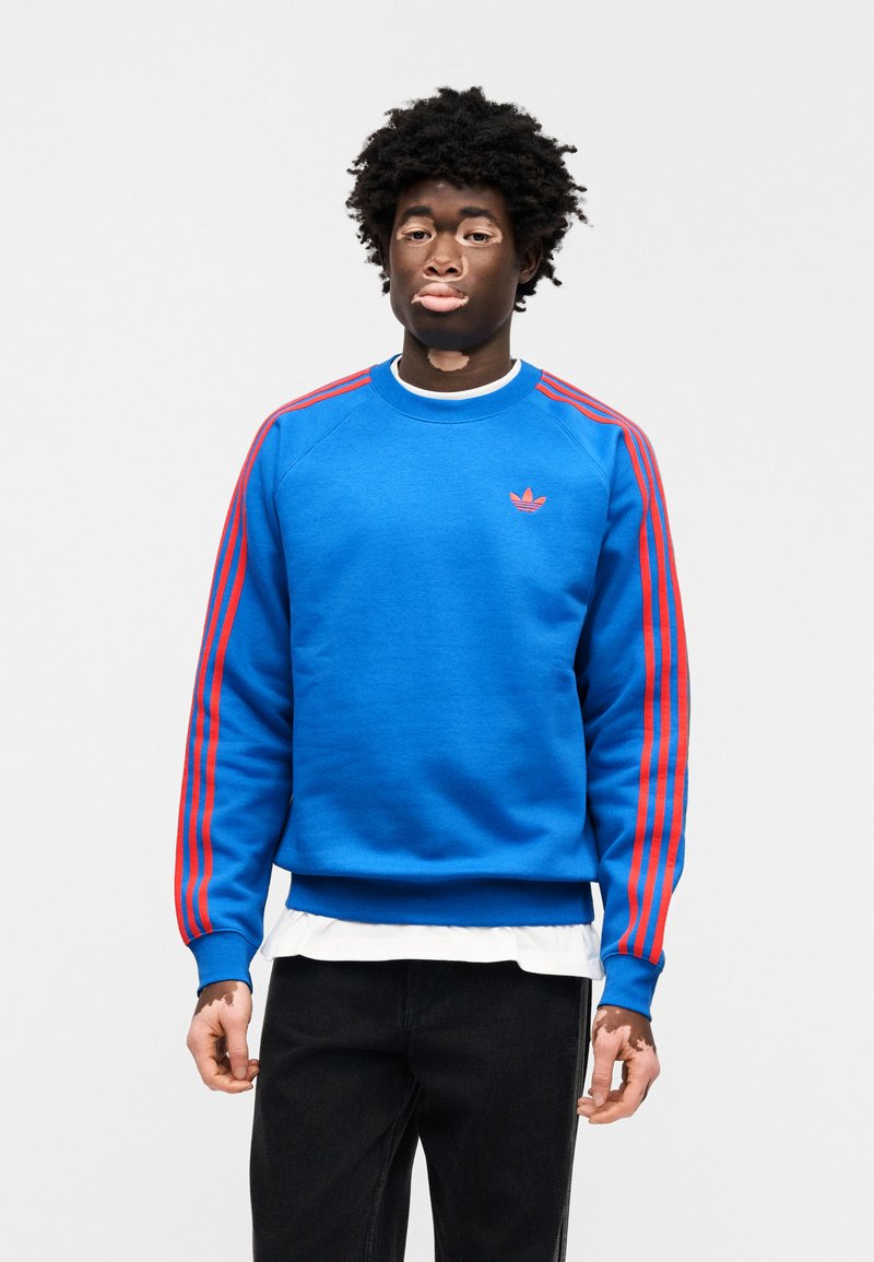 Sweat-shirt bleu en tissu doux, avec des rayures rouges sur les manches et un petit logo rouge sur la poitrine. Col rond.