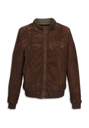 Homme debout les mains dans les poches portant une veste bomber vert olive, chemise blanche, pantalon large noir et chaussures noires.