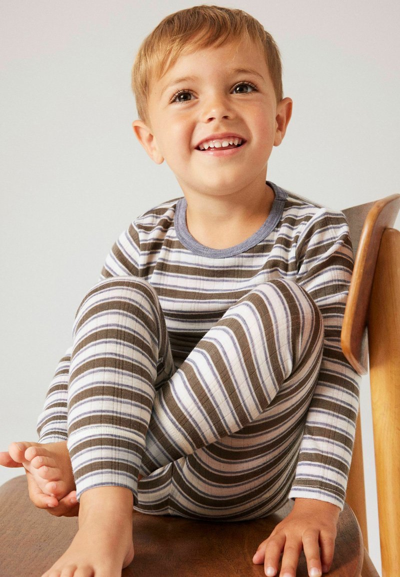 Jeune enfant souriant assis en tailleur sur une chaise en bois, portant un haut à manches longues rayé et un pantalon assorti dans des tons neutres.