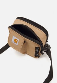 Tan crossover taske med dobbelte lynlåse, stoftekstur, frontlomme og et Carhartt logo patch. Sort justerbar rem fastgjort.