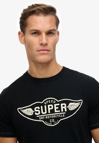 Zwarte katoenen t-shirt met een grote crèmekleurige grafische ontwerp met vleugels en tekst: "SUPER SPEED DRY MOTORCYCLE CO." rondom het logo.