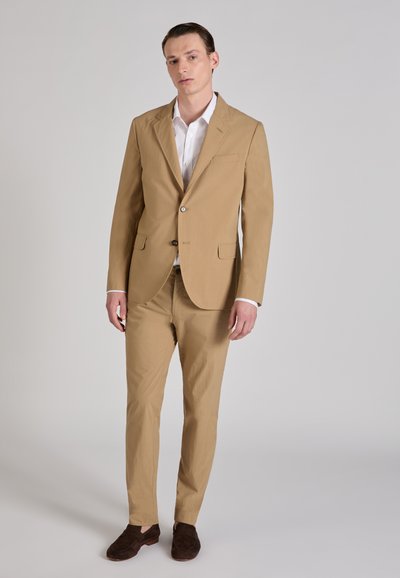 Beige pak met getailleerd jasje, twee voorzakken en sluiting met één knoop, gecombineerd met bijpassende broek en bruine suède schoenen.