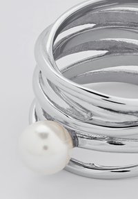 Silberfarbener Ring mit mehreren verdrehten Bändern und einem auffälligen weißen Perlenakzent. Glatte, glänzende Oberfläche mit strukturierten Akzenten.