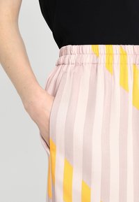 Růžové pruhované kalhoty s elastickým pasem a žlutými geometrickými akcenty. Viditelná boční kapsa, látka se zdá být lehká a mírně texturovaná.