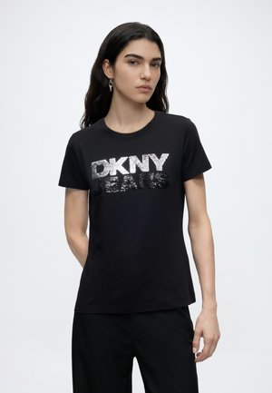 Femme aux longs cheveux foncés portant un t-shirt noir avec un texte en sequins argentés et noirs indiquant "DKNY JEANS" sur un fond gris clair.