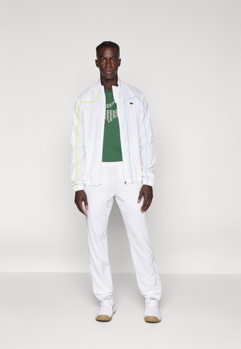 Biała kurtka z niebieskimi i żółtymi akcentami, połączona z zieloną koszulką z geometrycznym wzorem oraz białymi spodniami. Lekka tkanina, sportowy krój.