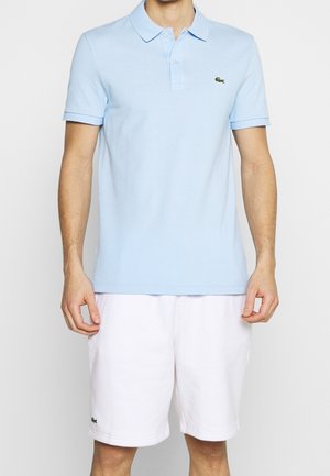Homme portant un polo bleu clair à manches courtes et un short blanc mi-genoux, debout devant un fond clair uni.