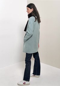 Cappotto verde chiaro a un petto con tasche laterali, indossato sopra jeans scuri e sneakers bianche. Sciarpa nera drappeggiata attorno al collo.