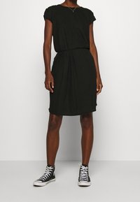 Schwarzes Kurzarmkleid mit lockerer Passform, geraffter Taille, kniefreier Saum, kombiniert mit schwarzen High-Top-Sneakern. Schlichte Stofftextur.