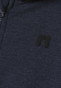 Felpa con zip di un blu scuro navy, con una texture morbida e un grande patch logo nero sul petto. Il materiale sembra traspirante e informale.