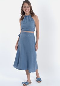 Top halter di un azzurro chiaro con scollatura arricciata abbinato a una gonna avvolgente dello stesso colore. Tessuto morbido e strutturato; sandali slip-on con fiocchi.