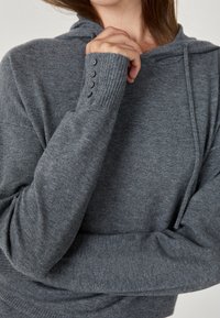 Frau trägt einen grauen Strickpullover mit Kapuze und vier Knöpfen am Ärmel, hält den Kragen mit der Hand nahe am Hals.