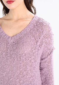 Fialový pletený svetr s výstřihem do V, volným střihem a otevřeným pletením. Texturovaná tkanina s jemným vzhledem a subtilními detaily na linii výstřihu.