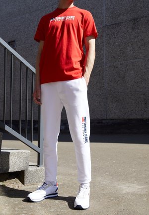 Person trägt rotes Tommy Jeans T-Shirt und weiße Jogginghose mit Tommy Jeans Logo, steht draußen nahe einer Steinmauer und Treppe.